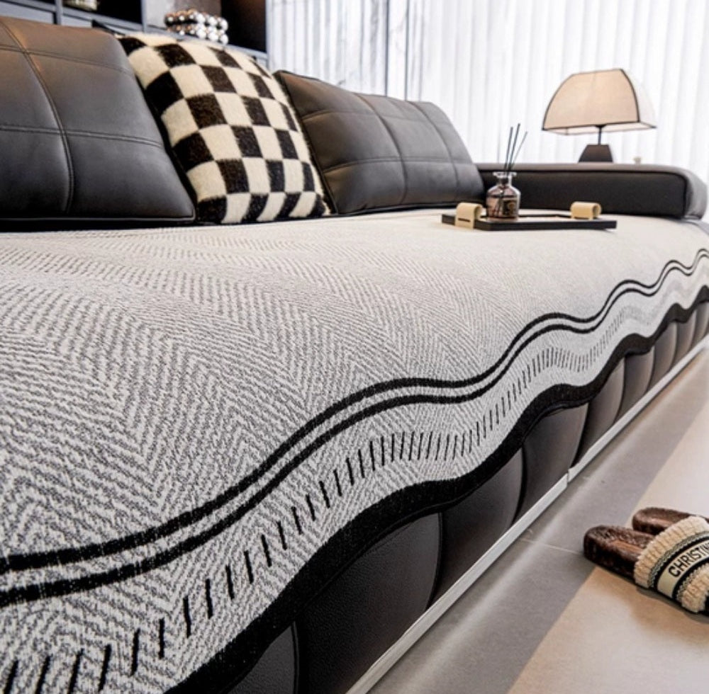 Sofa covered with a light gray woven blanket featuring a black wavy edge, black leather cushions, a black and white checkered pillow, a tray with decorative items, and slippers on the floor.