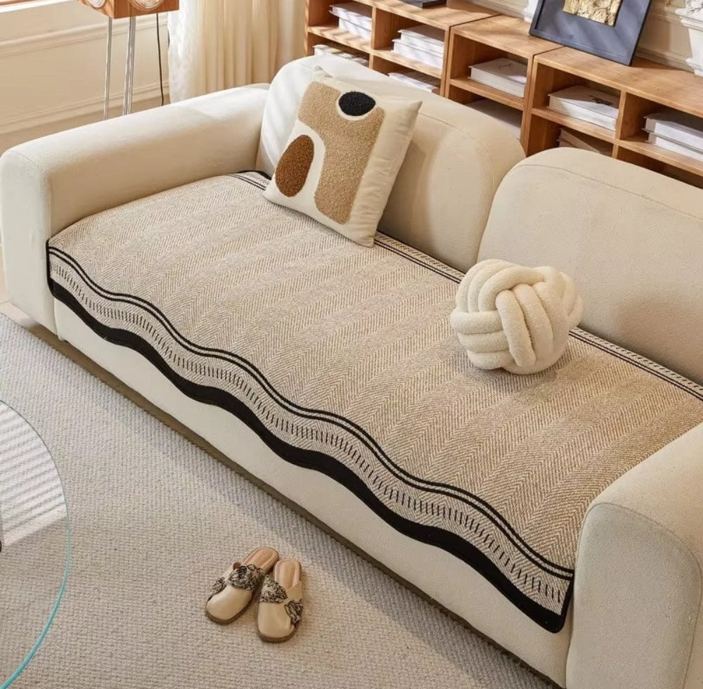 Cream sofa with a beige Seabrook sofa cover featuring a black wavy border, a decorative beige pillow, a knotted cream cushion, and beige slippers on a light rug in a living room with wooden shelves