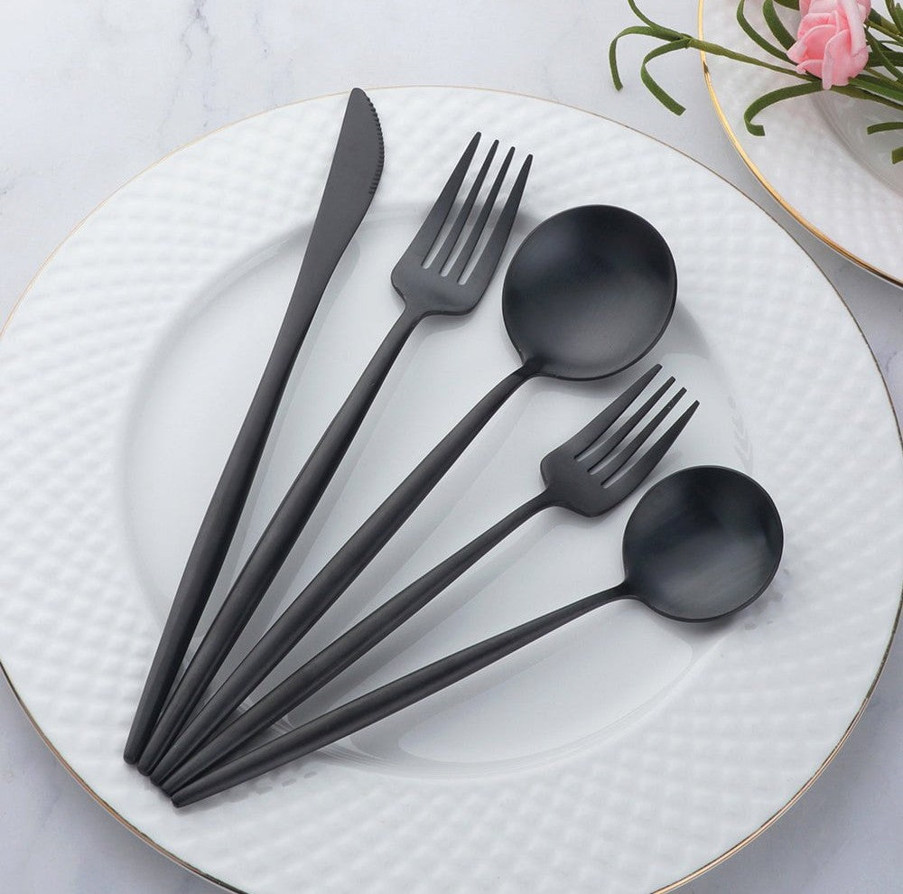 Black matte stainless steel cutlery set with a knife two forks and two spoons displayed on a white textured plate with a gold rim