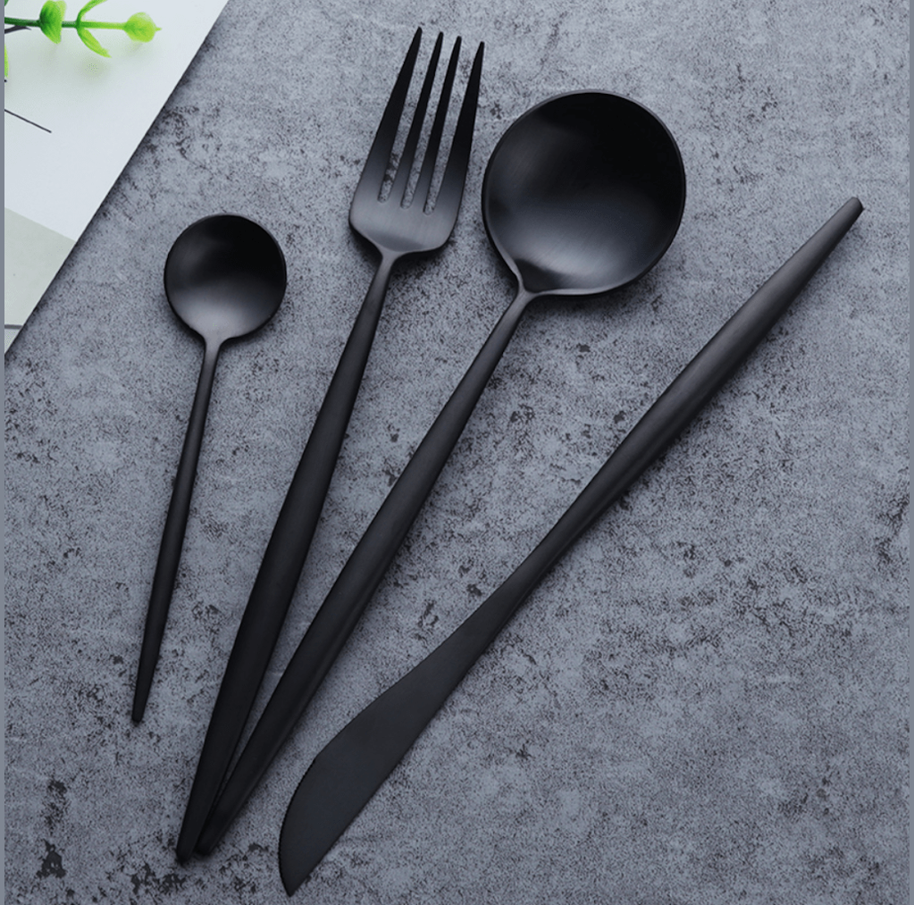 Black matte stainless steel cutlery set with slim curved handles including a small spoon a fork a large spoon and a knife laid on a gray textured surface