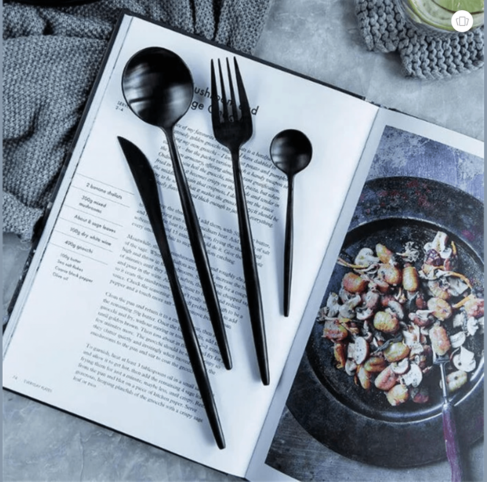 Black stainless steel cutlery with slim curved handles laid across an open magazine featuring a large spoon a fork a knife and a small spoon beside a plate of food