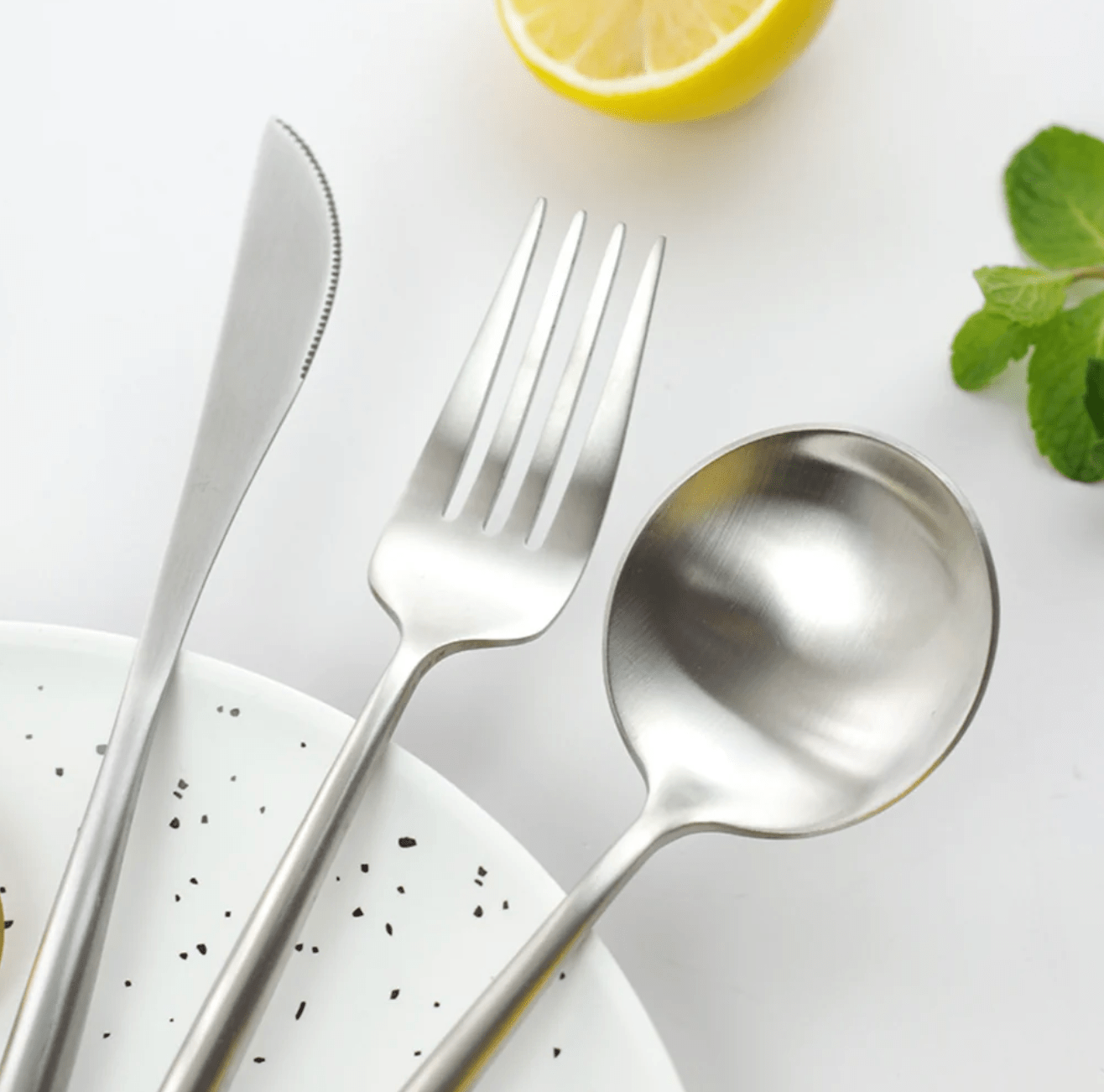 Stainless steel cutlery set with slim curved handles including a knife a fork and a spoon arranged over a white speckled plate with a lemon half and mint leaves