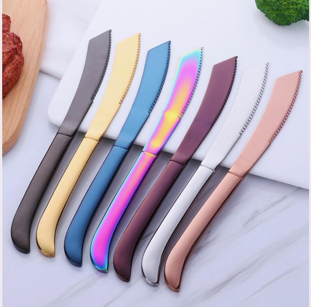 A row of colorful stainless steel kitchen knives with serrated blades and a rainbow iridescent blade in the middle laid on a white surface beside a wooden cutting board