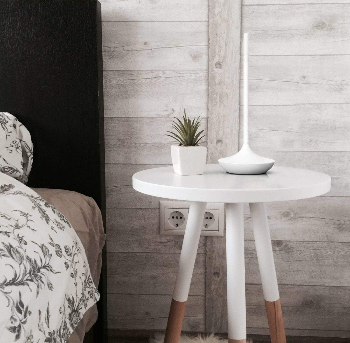 Round white side table with a small square plant pot and a tall white lamp against a wood paneled wall beside a bed with floral bedding and an electrical outlet visible