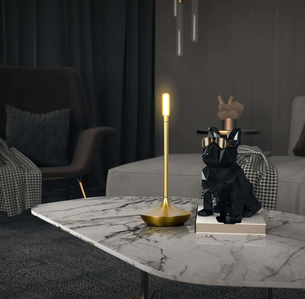 A lit gold brass LED table lamp with a slender stem and round base sits on a white marble coffee table beside a black geometric bulldog statue with a gold bow tie in a dark living room.