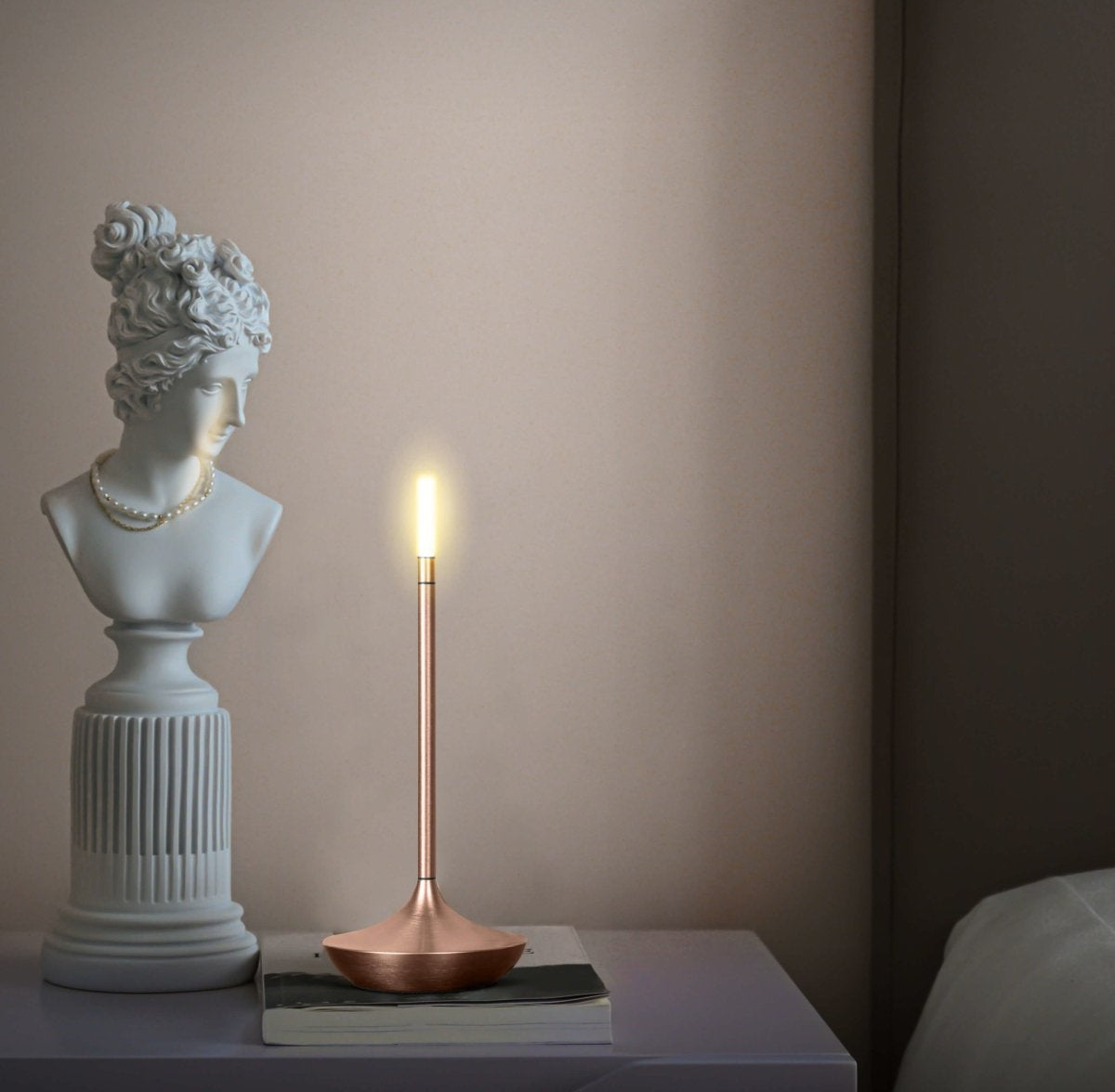 Slender copper table lamp with a vertical LED bulb glows softly on a white table next to a classical bust statue wearing a pearl necklace.