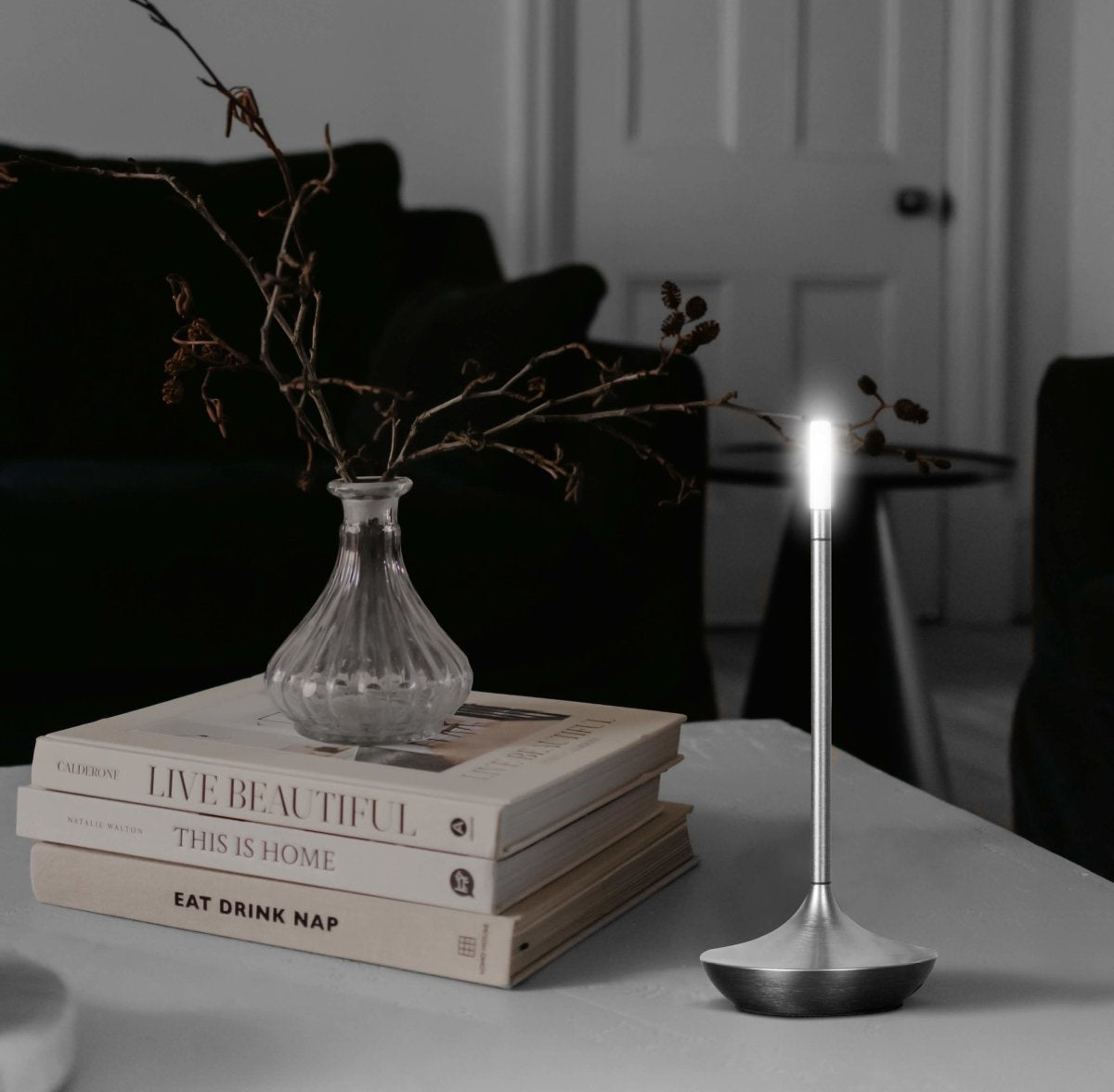 A modern silver LED table lamp with a slender stem and round base glows on a white table beside a stack of books and a glass vase with dried branches.