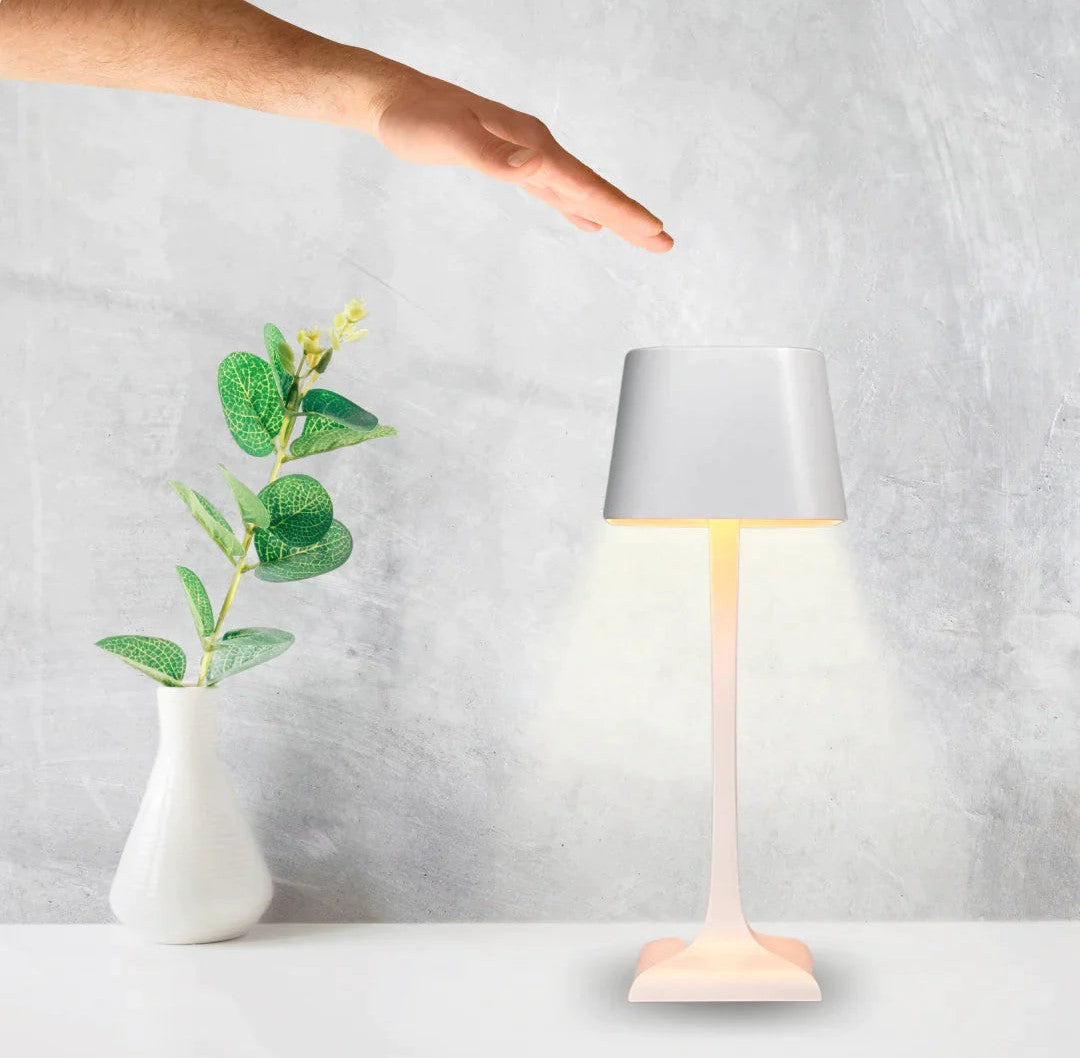 A pink cordless table lamp with a gray shade on a white surface beside a white vase with green leafy plant, with a hand hovering above the lamp.