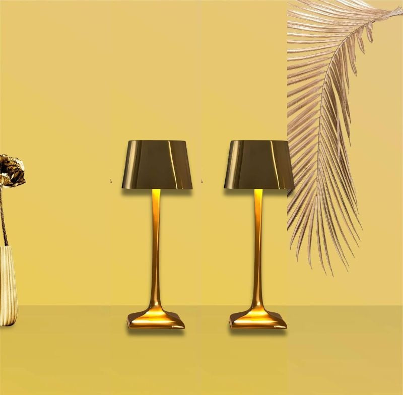 Two gold table lamps with black shades on a yellow surface, a vase with a dried flower on the left and a palm frond on the right