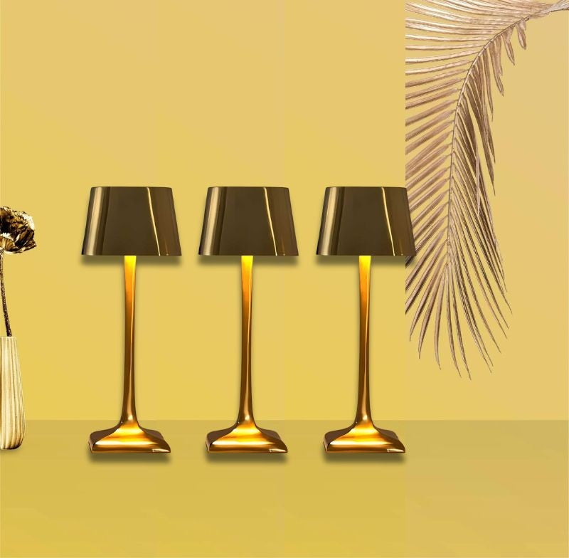 Three gold cordless table lamps with black shades on a yellow background, with a vase on the left and a palm frond on the right