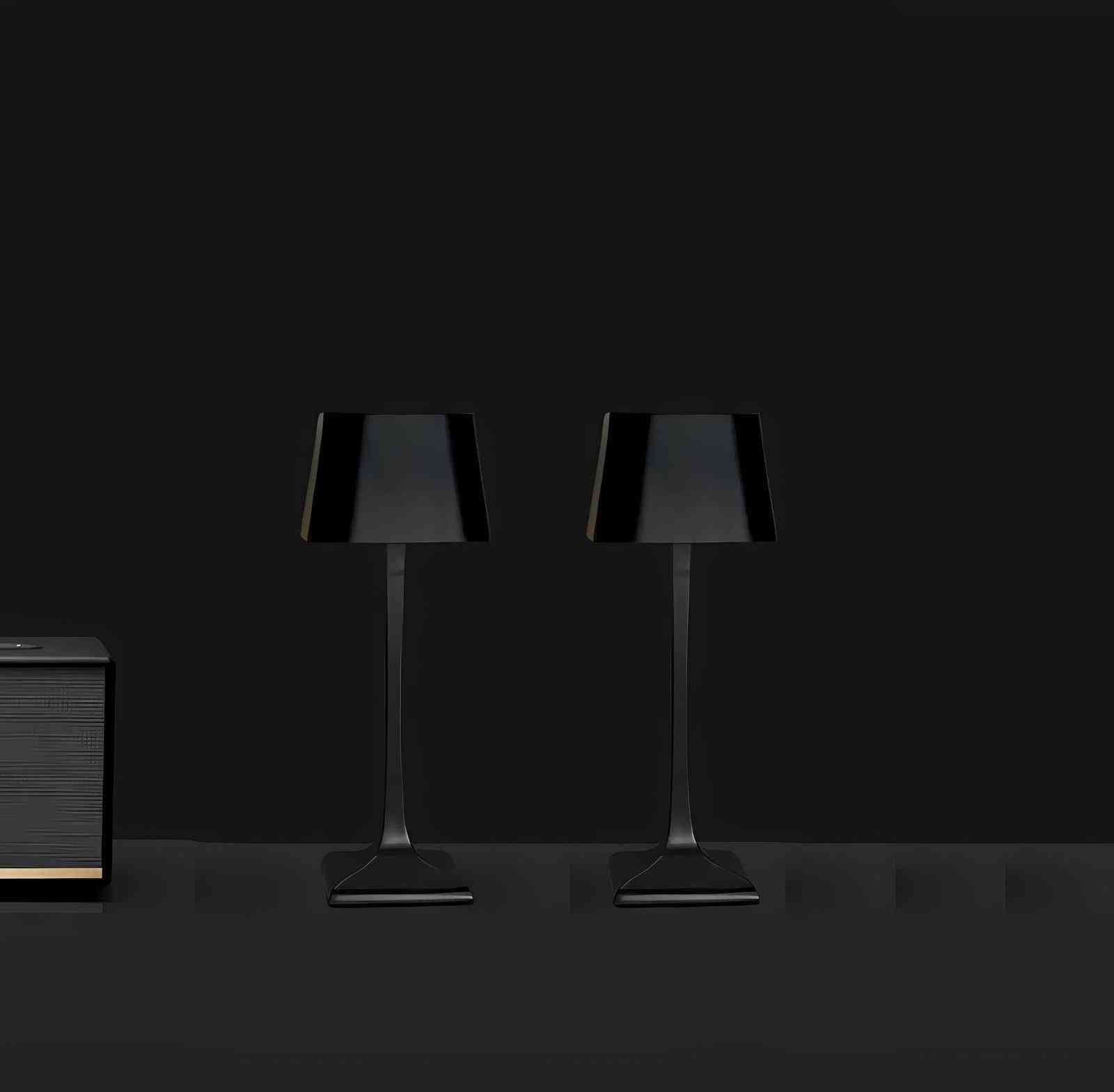 Two black cordless table lamps with square shades and slim stems sit on a dark surface, with a small wooden cabinet on the left.