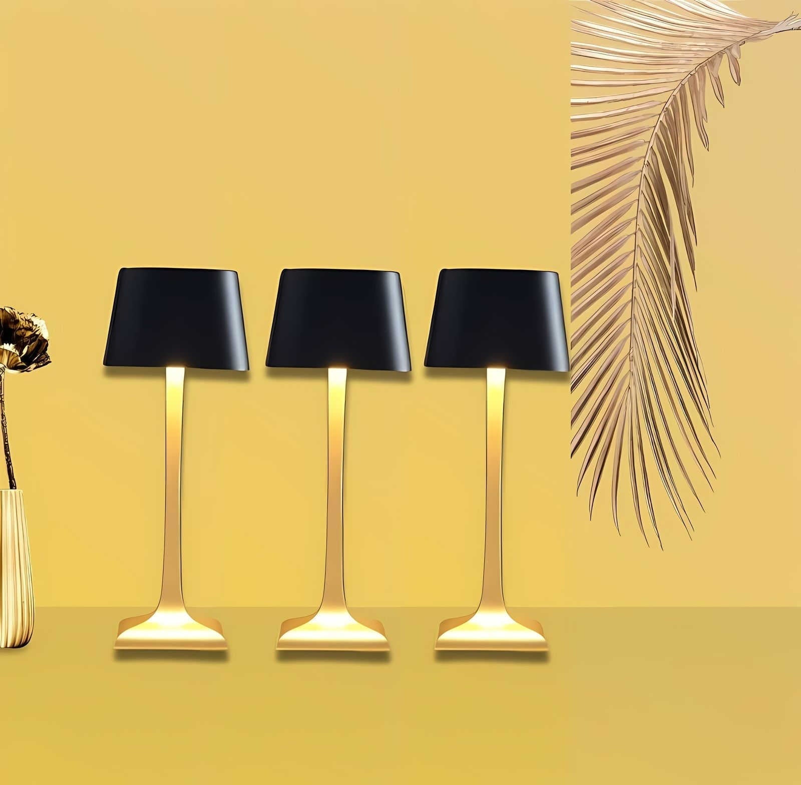 Three cordless table lamps with black shades and gold metal stems on a yellow background, with a vase on the left and a palm leaf on the right