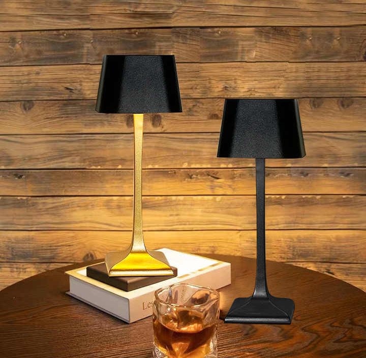 Two cordless table lamps with black shades and metal stems on a wooden table, gold lamp on the left and black lamp on the right, with a glass of whiskey and stacked books nearby.