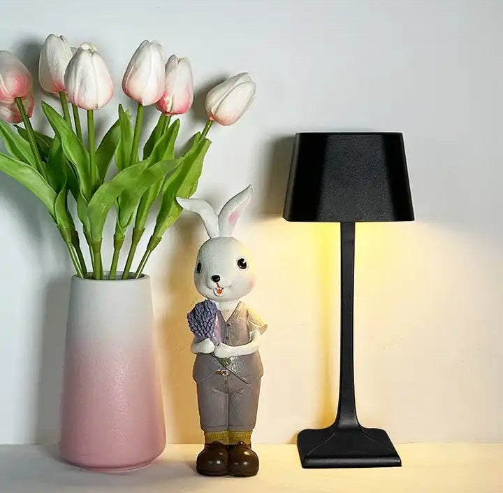 A vase of pink tulips beside a rabbit figurine and a black cordless table lamp with a square shade on a light wall.