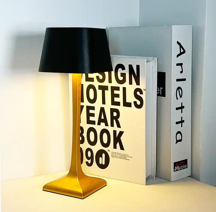 Gold metal cordless table lamp with a black fabric shade sits on a white surface beside stacked white books with bold black text