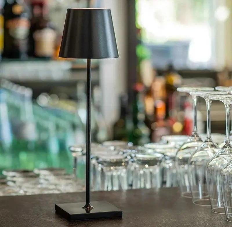 Black cordless table lamp with a slim stem and square base on a bar counter, black lampshade with glassware in the background