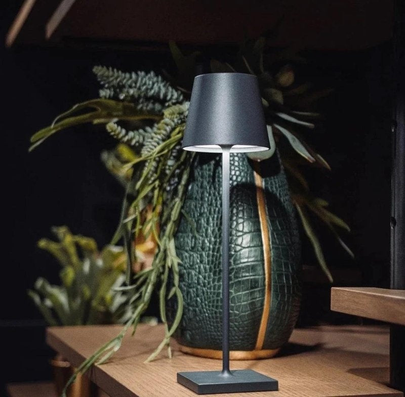 Cordless table lamp with a dark gray shade, slim metal stem, and square base on a wooden surface, with a green textured vase and foliage in the background