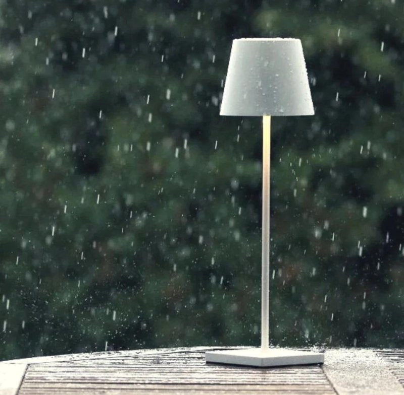 Cordless table lamp with a powder coated aluminium body and a light gray shade on a square base sits on a wooden surface in the rain.