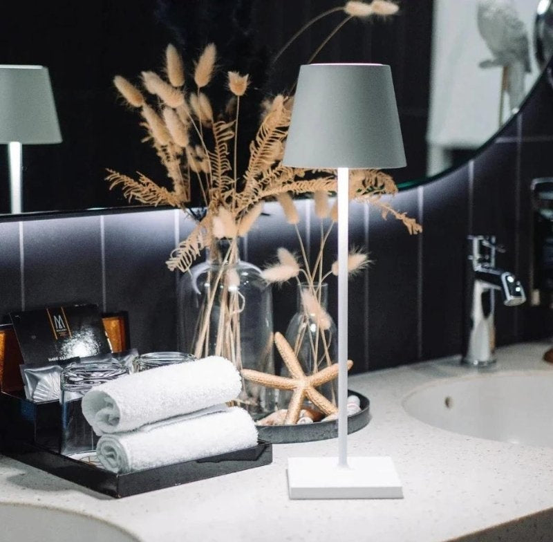 Cordless white table lamp with a gray lampshade and square base on a speckled bathroom counter, beside rolled towels and decorative dried plants with a starfish in a glass vase