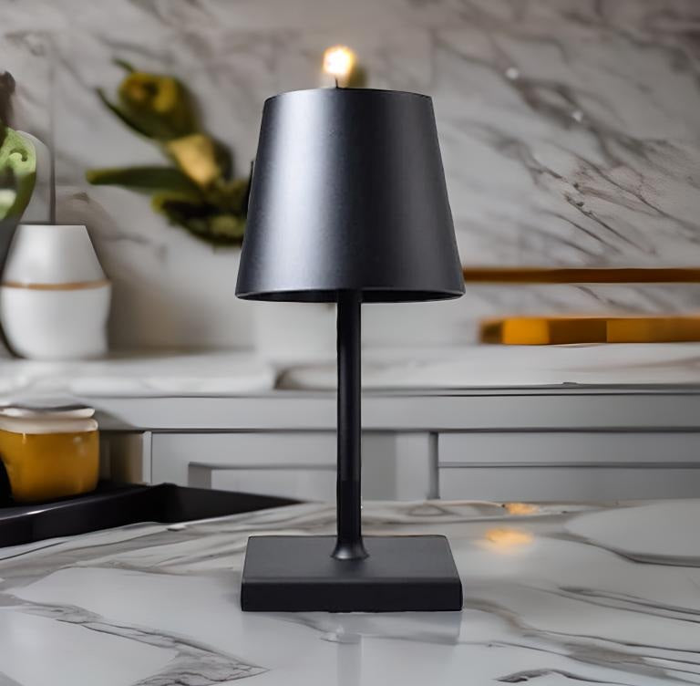 Black metal table lamp with a slim stem and square base, a black shade, and a small warm light at the top on a marble countertop.
