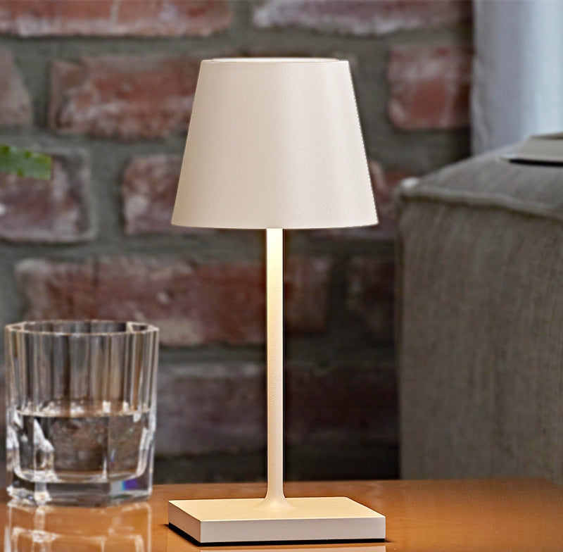 Beige table lamp with a cream tapered shade and slim stem on a square base set on a wooden table beside a glass of water and a brick wall background