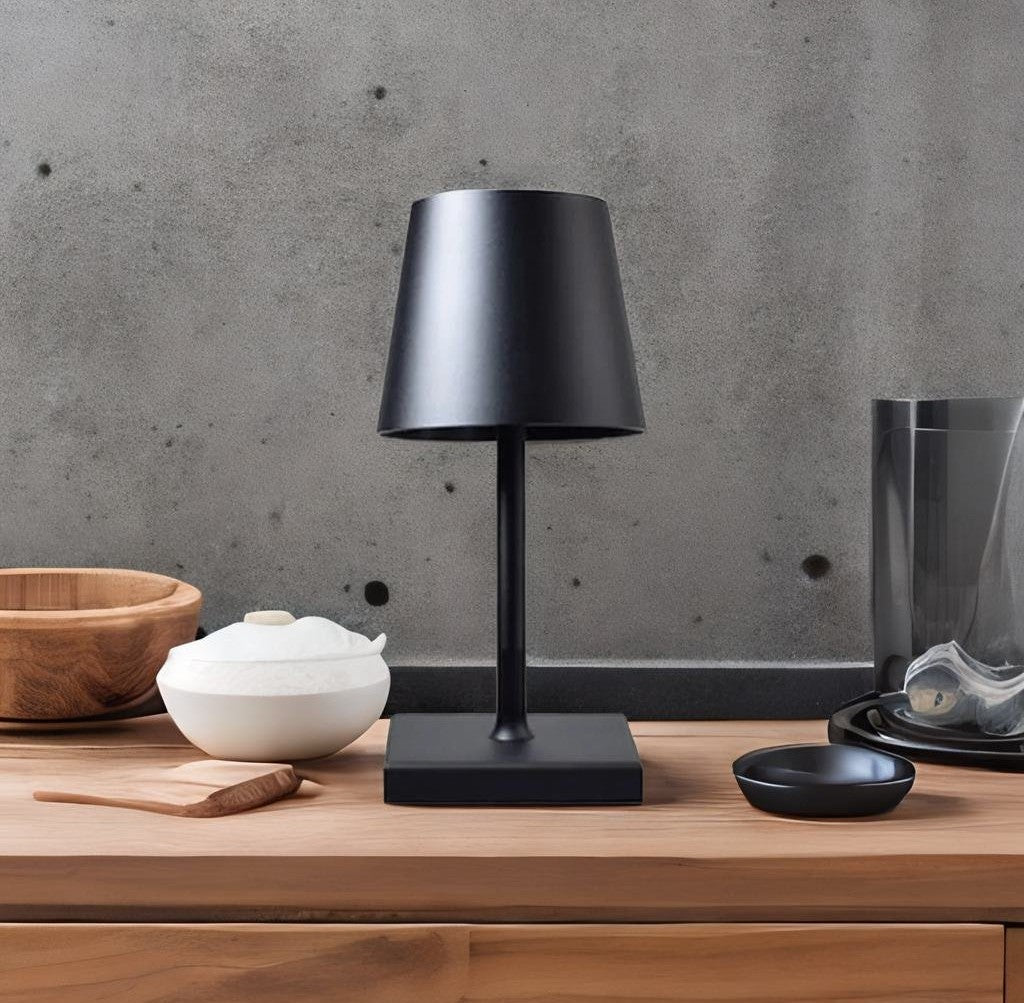 Black touch sensor table lamp with a conical shade on a square base sits on a wooden surface, with a wooden bowl and a white lidded bowl to the left and a glass cloche to the right against a gray concrete wall