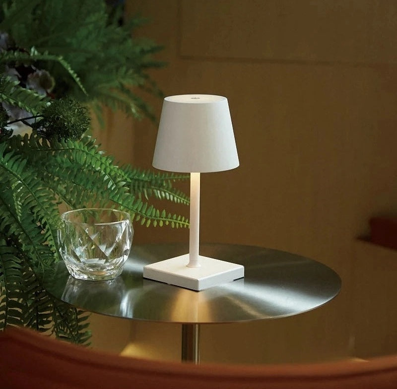 White table lamp with a square base and conical shade on a round metal table beside a crystal glass and green ferns