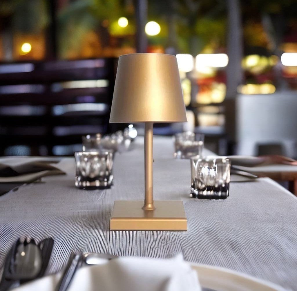 Gold metal table lamp with square base and slender stem on a striped dining table with glassware in the background