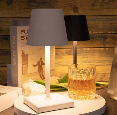 Two table lamps with gray and black shades on a white round base on a wooden surface, with a textured amber glass tumbler and books in the background.