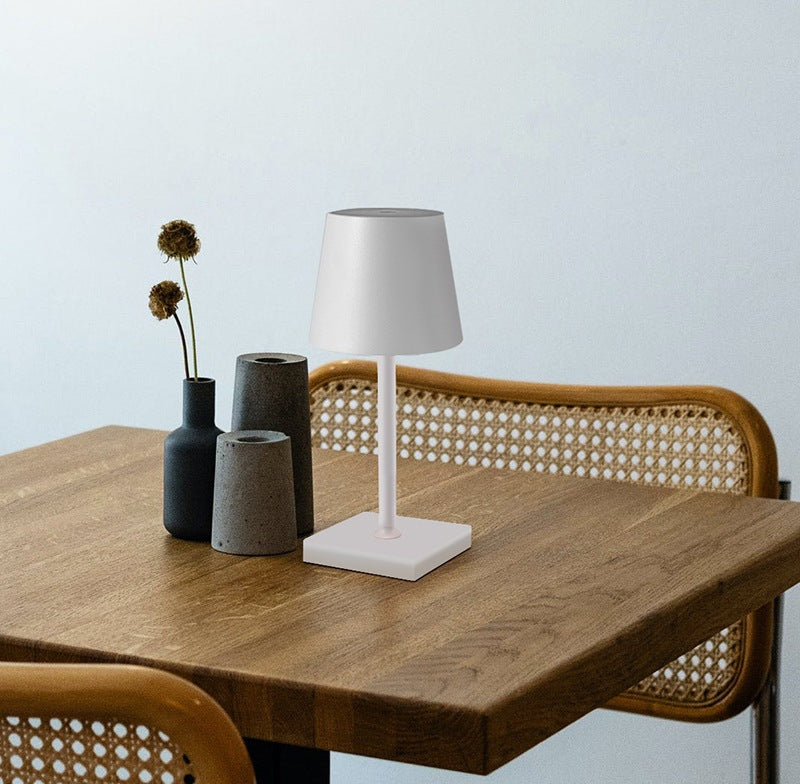 White table lamp with a conical shade on a slender white stem and square base sits on a wooden table beside a group of three dark vases and a woven back chair