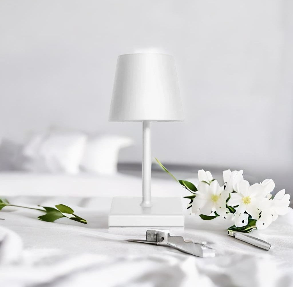 White table lamp with shade and square base on a white surface, with white flowers and metal tools in the foreground