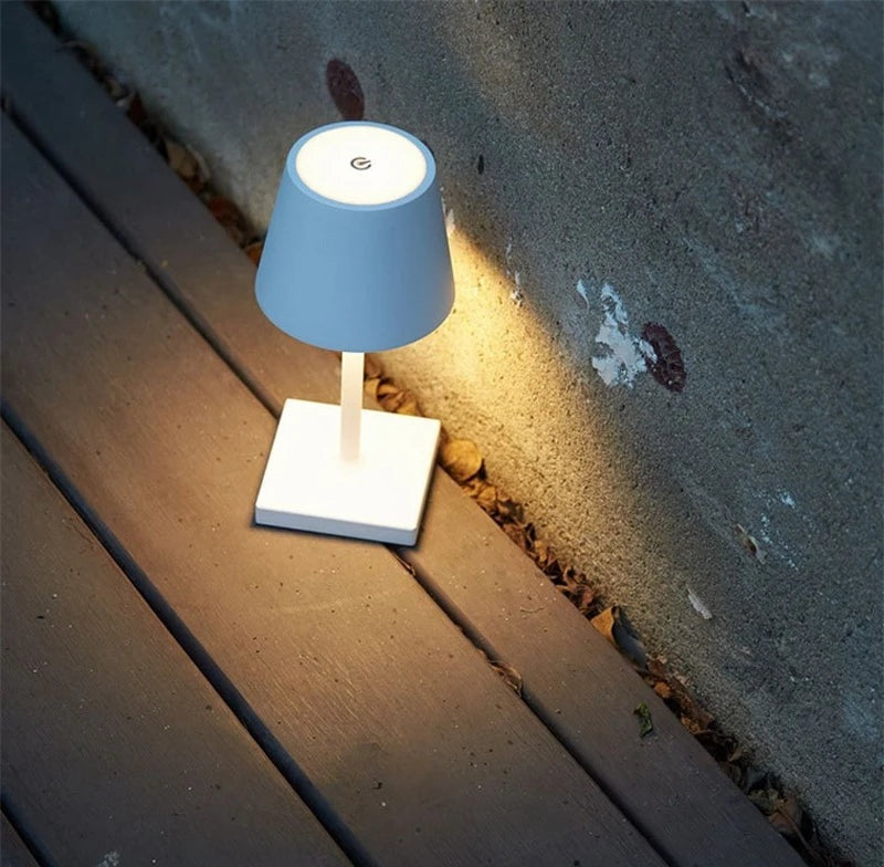 Blue table lamp with a white square base and white stem on a wooden deck, blue shade with a round touch sensor on top, casting warm light on a concrete wall.