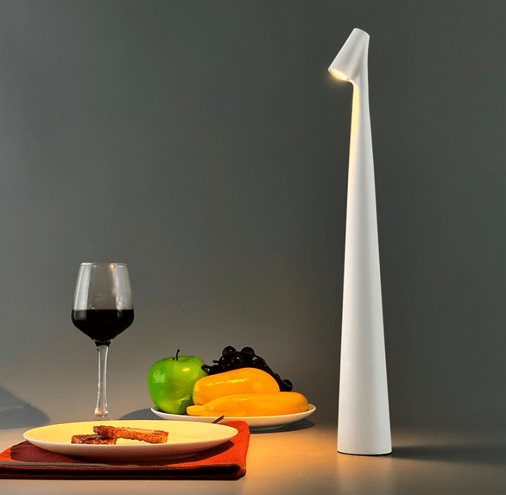Tall white wireless touch control table lamp with a slim tapered base and warm downlight on the right, beside a dining setup with a wine glass and plates of crackers and green apple, yellow peppers, and grapes on a red placemat.