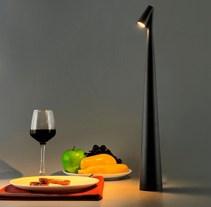 Tall black aluminum wireless touch control table lamp with a slim curved stem and an angled lamp head casting warm light over a table setting with a wine glass and plates of fruit and bread.