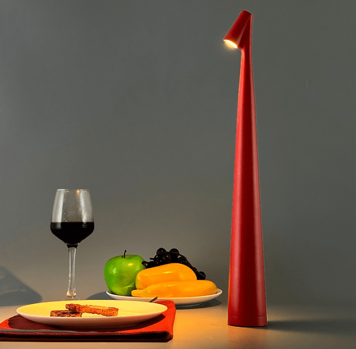 A tall red slim table lamp with a small lit head stands on a surface beside a glass of red wine, a green apple, a plate of yellow fruit and black grapes, and a plate with toast on a red napkin.