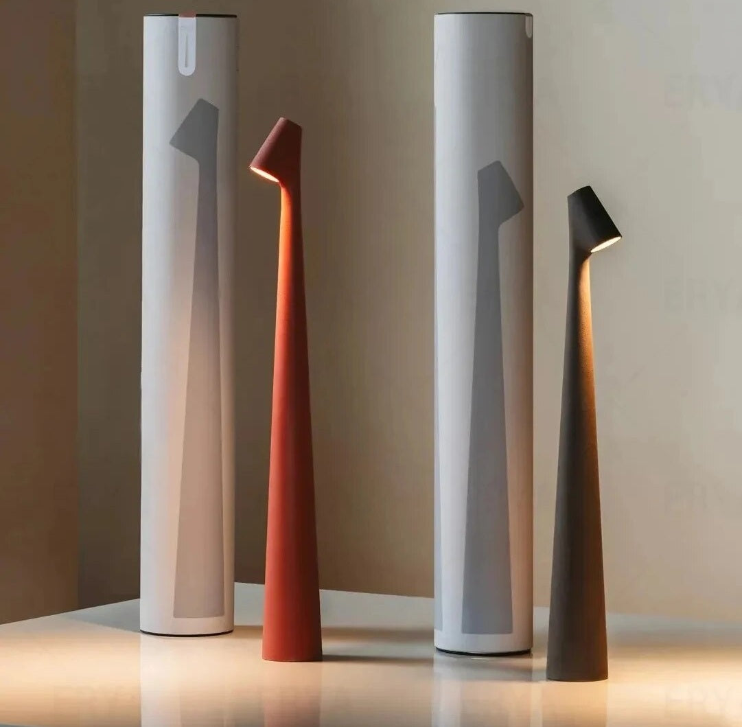 Four modern lamps on a light wooden surface, two tall white cylindrical bases in the back and two slim orange and dark gray cone shaped lamps in the front with angled heads.