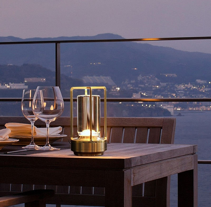 gold metal framed lantern with glass cylinder on a wooden outdoor dining table beside wine glasses and plates with a dusk harbor and hills in the background