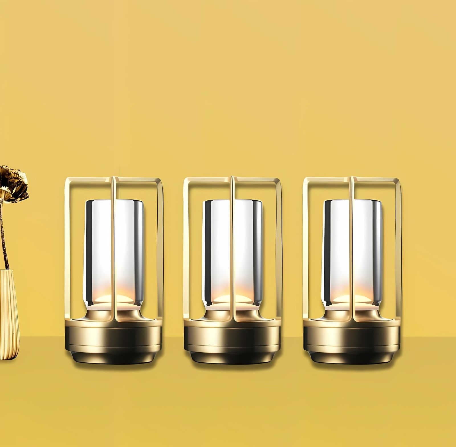 Three gold cordless lantern table lamps with crystal glass cylinders and metal frames on a yellow background, with a vase of dried flowers on the left.