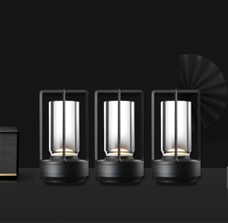 Three cordless lantern table lamps with crystal glass cylinders inside black metal frames glowing warm light on a dark surface