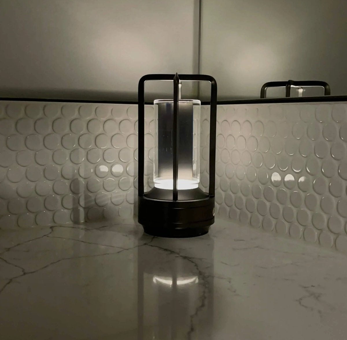 Cordless lantern style table lamp with a metal frame around a glass cylinder illuminated on a white marble countertop with a bubble tile backsplash