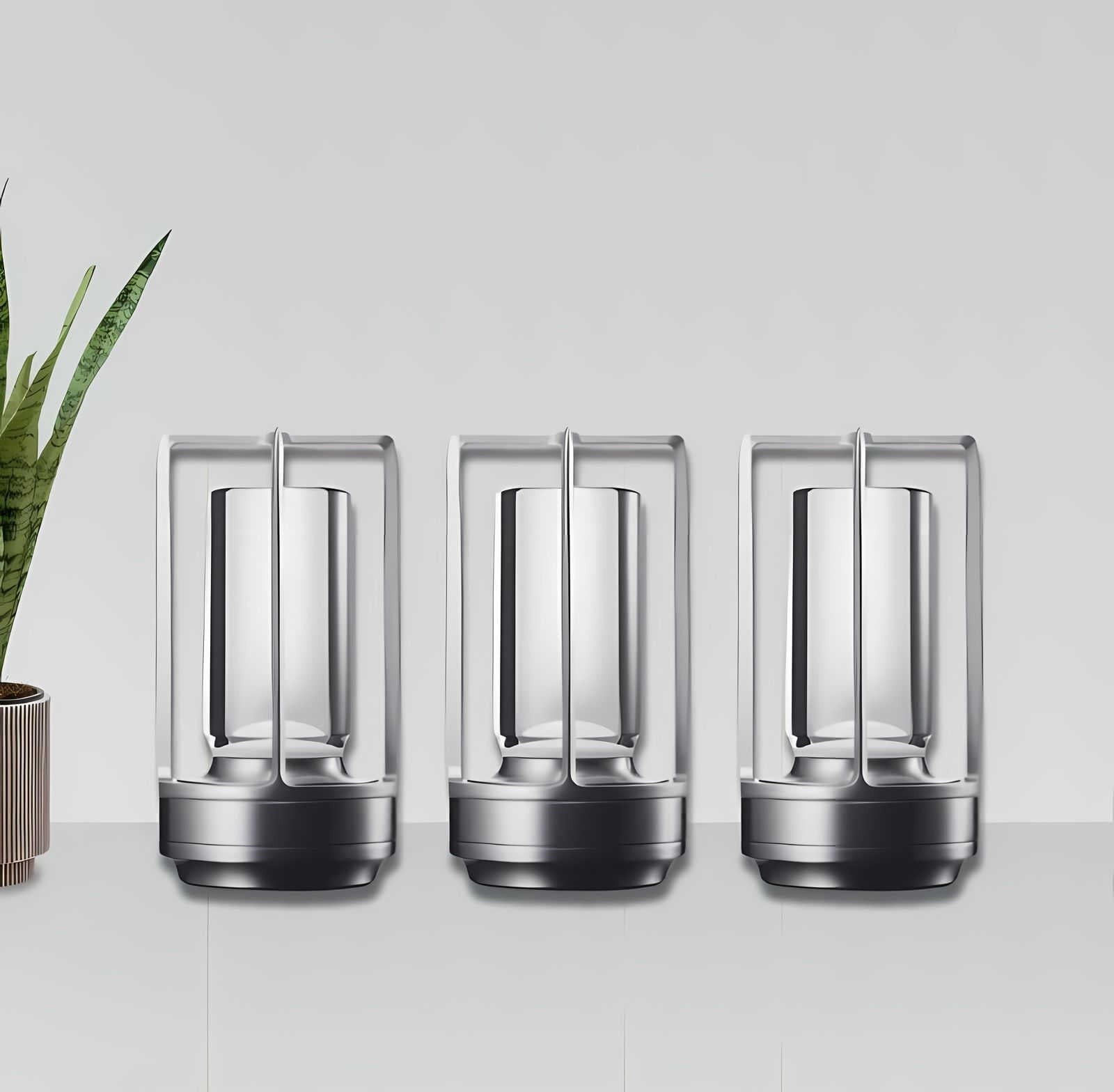 Three silver cordless lantern table lamps with metal frames and glass panels lined up on a white surface with a potted plant to the left