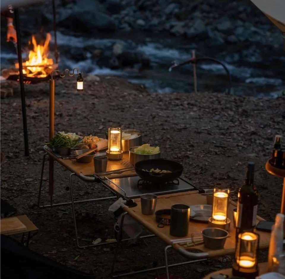 Outdoor camping scene showing cordless lanterns with warm light on a wooden folding table set with plates cups and bottles fire pit glowing in the background