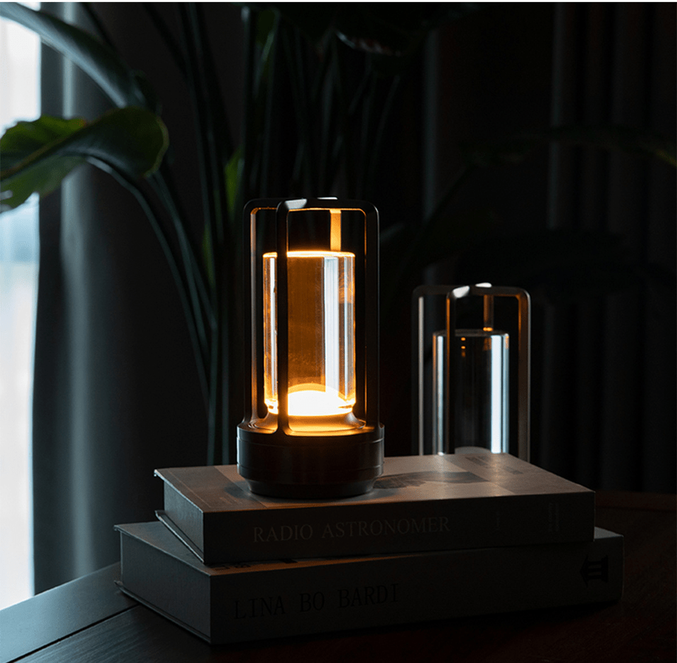 Cordless lantern table lamp with crystal panels inside a metal frame glowing warm amber on top of stacked books beside a smaller lantern on a dark wooden table