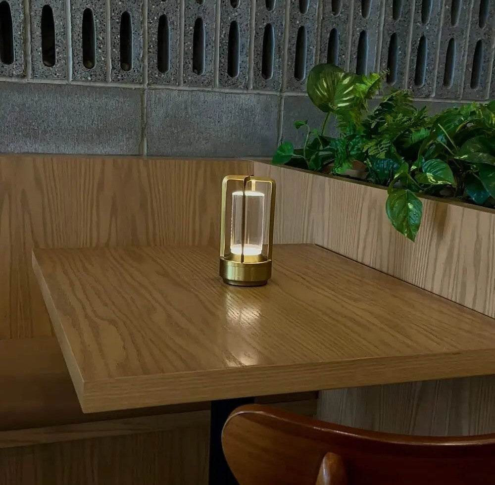 Gold framed cordless lantern table lamp with a glass crystal center on a wooden table with green plants in the background