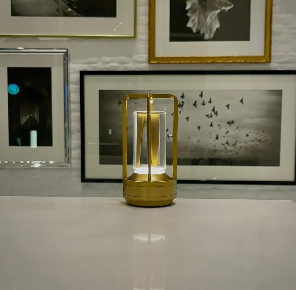 Gold cordless lantern table lamp with glass center glowing on a white tabletop with framed black and white photos on the wall behind