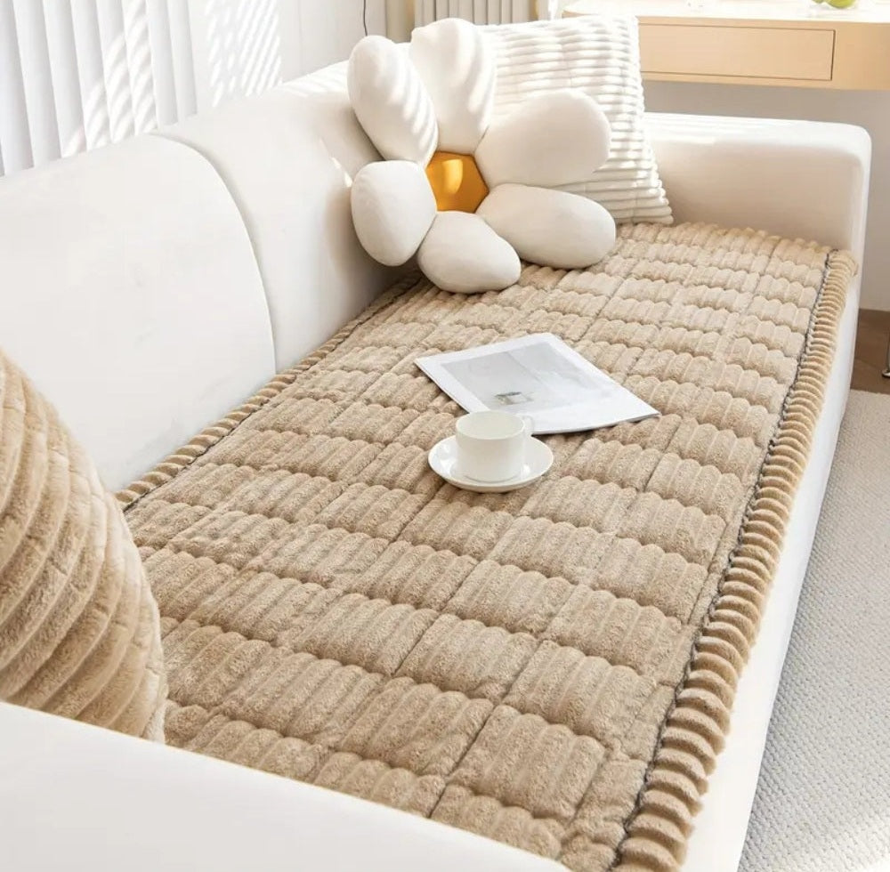 Beige microfiber sofa cover with plush flannel and hand pleated edges draped over a white sofa with a magazine and a white cup on top