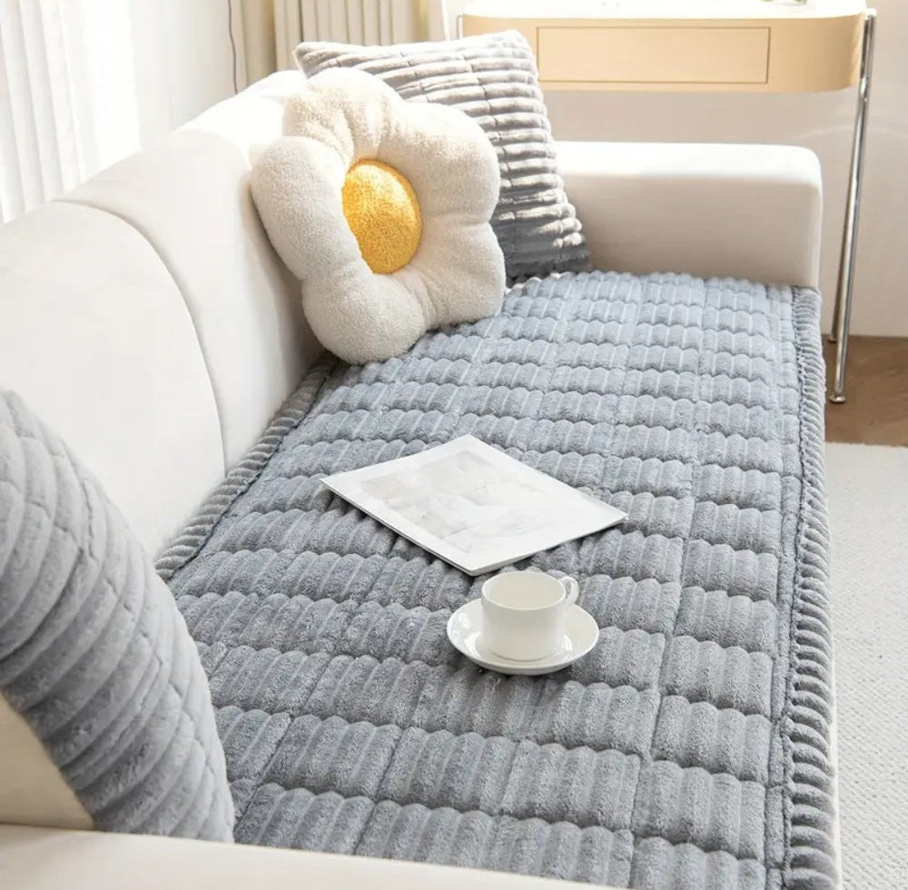 Veloura sofa cover in gray microfiber blend with plush flannel and hand pleated edges on a white sofa featuring a flower shaped plush pillow a gray striped cushion and a white cup on a saucer with a magazine