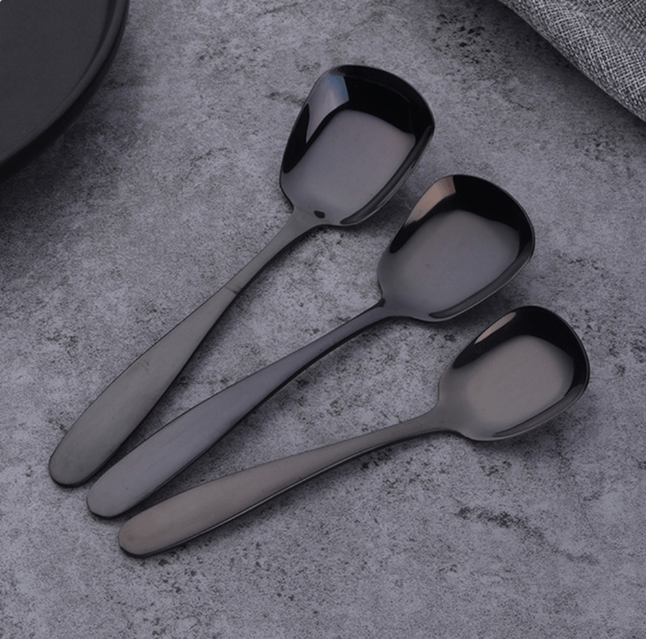 Three stainless steel serving spoons with a polished silver finish laid diagonally on a gray textured surface with a plate edge and a cloth in the background