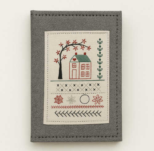 Grey fabric book cover with a beige embroidered patch featuring a house with a green roof under a tree and several decorative stitch patterns